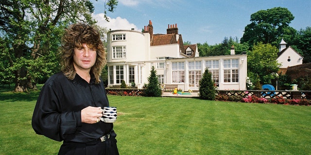 Black Sabbath singer Ozzy Osbourne holding a cup of tea in the garden of his home while wearing a black robe