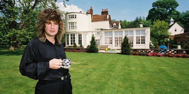 Black Sabbath singer Ozzy Osbourne holding a cup of tea in the garden of his home while wearing a black robe