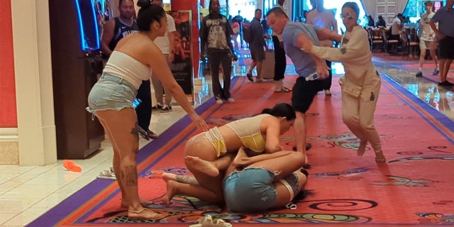 A woman in a thong is shown tussling with a woman in cut off shorts on the floor of a casino.