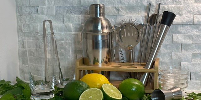 Barware kit with glasses, tongs, cocktail shaker, strainer, muddler spoons, and straws sit next to mojito ingredients.