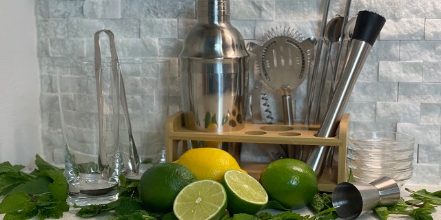 Barware kit with glasses, tongs, cocktail shaker, strainer, muddler spoons, and straws sit next to mojito ingredients.