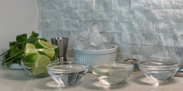 Mint sprigs on a coaster next to a container of ice and bowls of sliced limes, tequila, simple syrup and sparkling water.