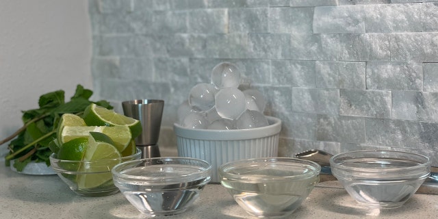 Mint sprigs on a coaster next to a container of ice and bowls of sliced limes, tequila, simple syrup and sparkling water.