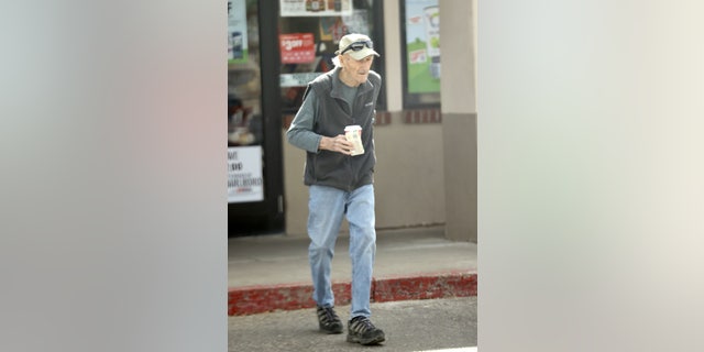 Gene Hackman crosses the street wearing baseball hat and sunglasses.