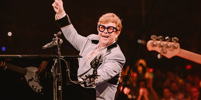 Elton John sitting at piano and pointing up