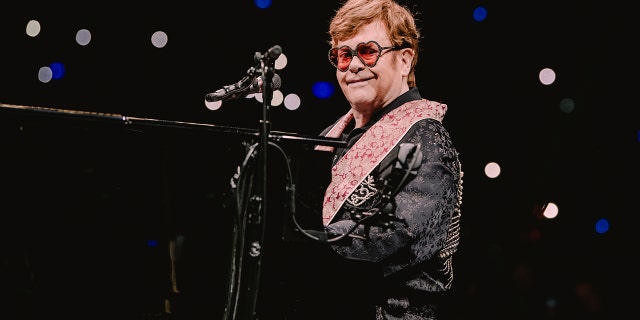 Elton John sits at piano smiling wearing sunglasses