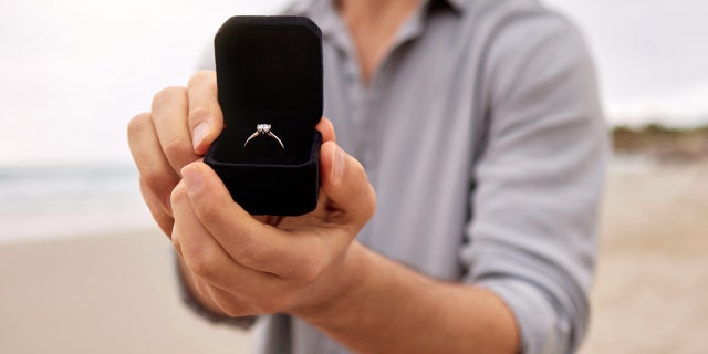 Shot of a man on a beach holding an open jewelry box with an engagement ring.