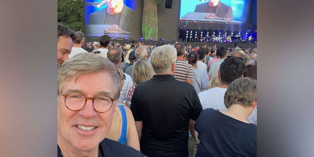 Greg Palkot lächelt im Selfie, während Billy Joel hinter ihm auftritt 