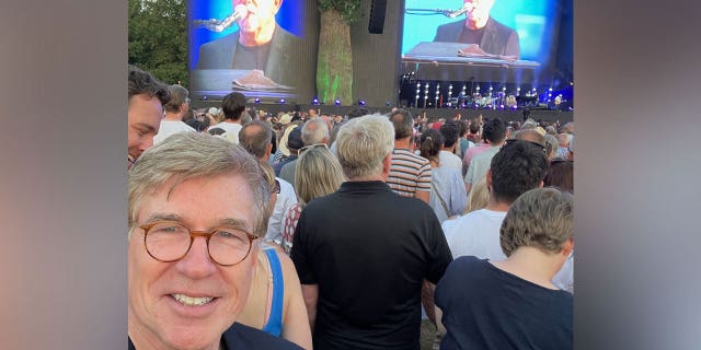 Greg Palkot smiles in selfie with Billy Joel performing behind him 