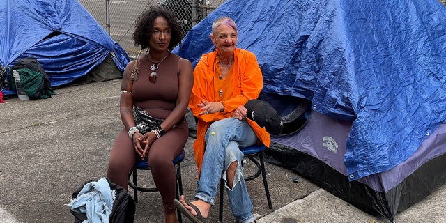 Two women sit on sidewalk in front of tents