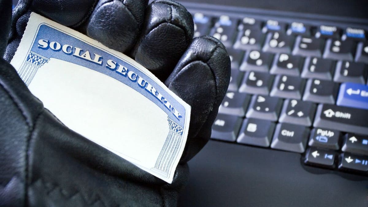 Picture of a cybercriminal holding a social security card in one hand while wearing a black glove and by a computer keyboard