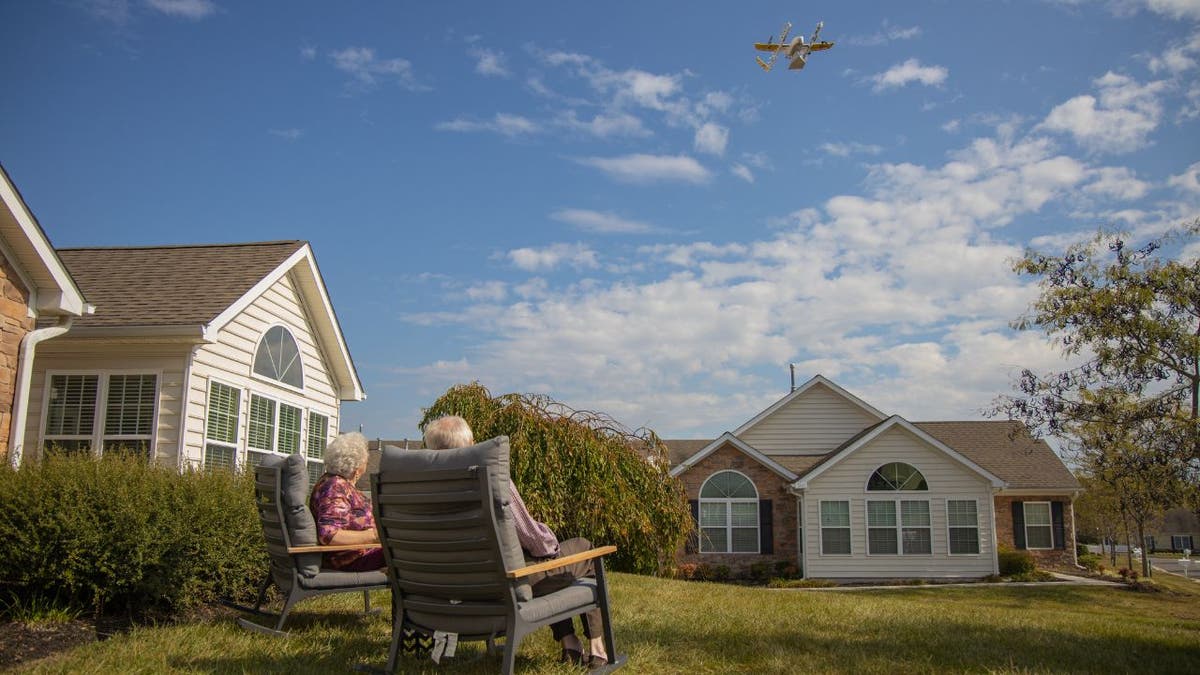 Susie and Paul sitting in chairs outside as a drone in the sky brings them their order