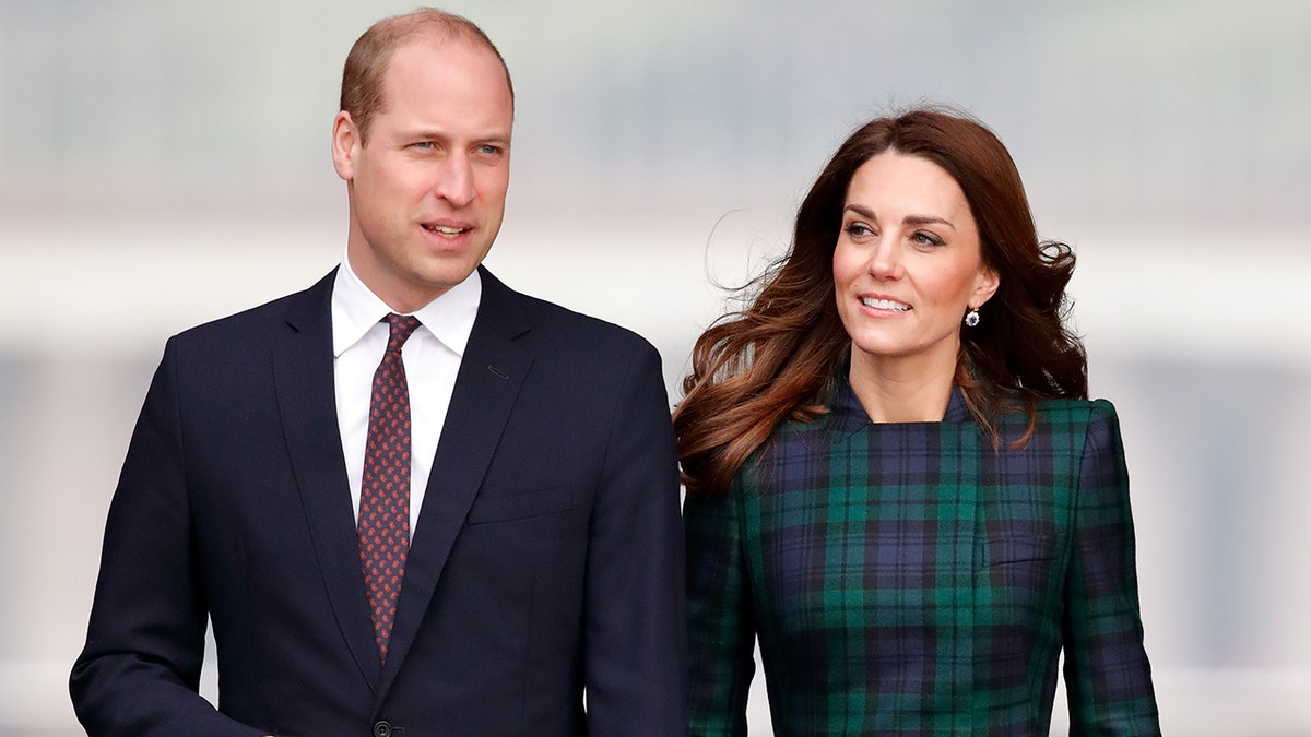 A close-up of Prince William wearing a dark blue blazer with a white shirt and red tie standing next to Kate Middleton in a green plaid dress