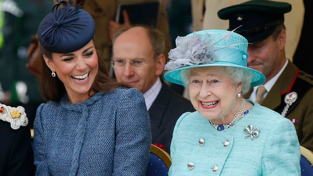 A close-up of Kate Middleton in a dark blue suit sitting next to a smiling queen elizabeth in a bright light blue suit