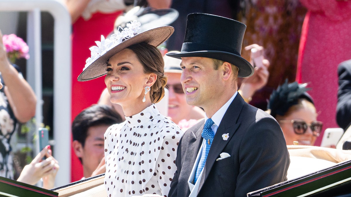 Kate Middleton in a black and white polka dress sitting next to Prince William in a suit and top hat