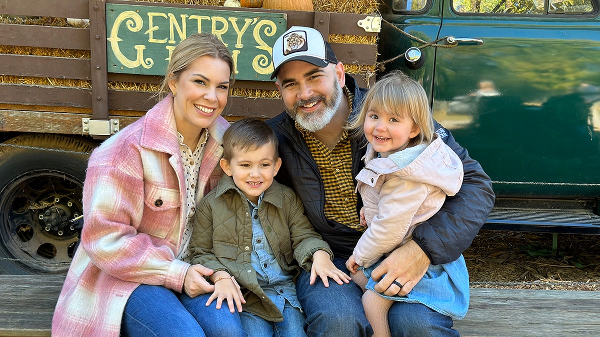 Tasha Layton wearing a pink plaid jacket, a printed blouse and jeans posing with her smiling husband and two children