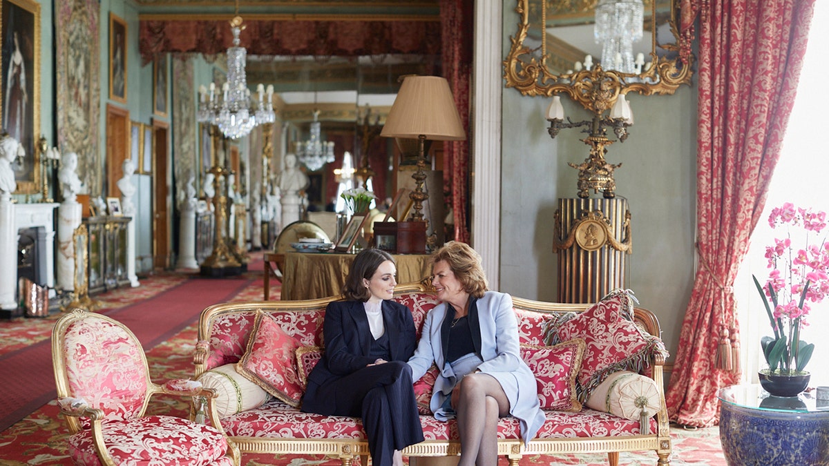 Lady Violet Manners in a navy blazer and pants with a white turtleneck sitting on a pink couch with her mother wearing a light blue blazer and a matching skirt.