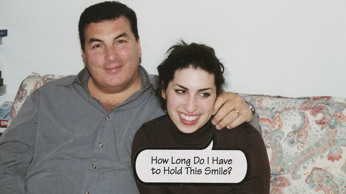Mitch Winehouse wearing a grey shirt putting his arm on Amy Winehouse who is wearing a black turtleneck