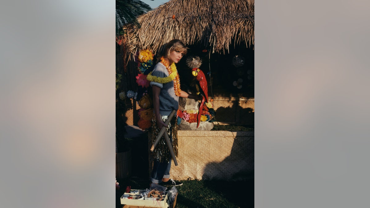 Michelle Pfeiffer wearing leis over a grey sweatshirt, with a gold-coloured grass skirt over a pair of jeans, holding a model of a red parrot while standing beside a thatched hut, on the set of Grease 2