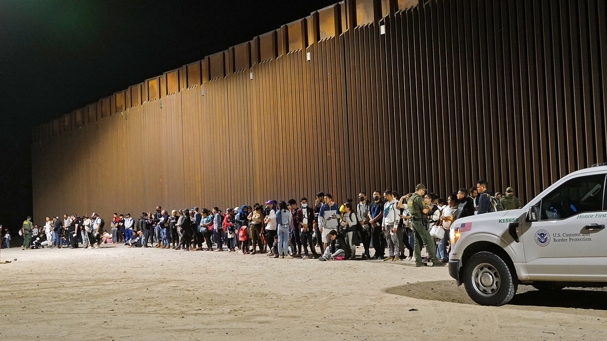 Migrants line up along the border wall.