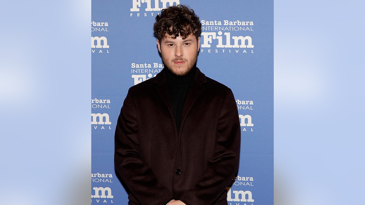 Nolan Gould in a turtleneck and jacket stares at the camera on the carpet