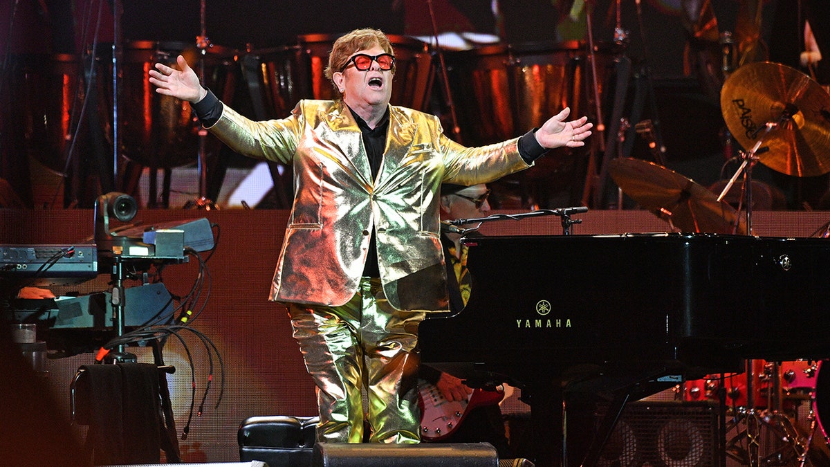 Elton John in a gold suit jacket and matching pants with his arms open on stage