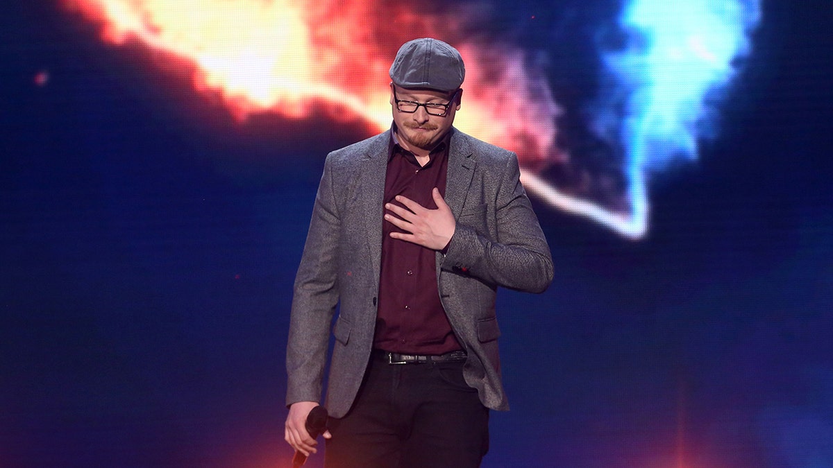 Art Garfunkel Jr. wearing a gray blazer and matching hat with a burgundy shirt performing on stage