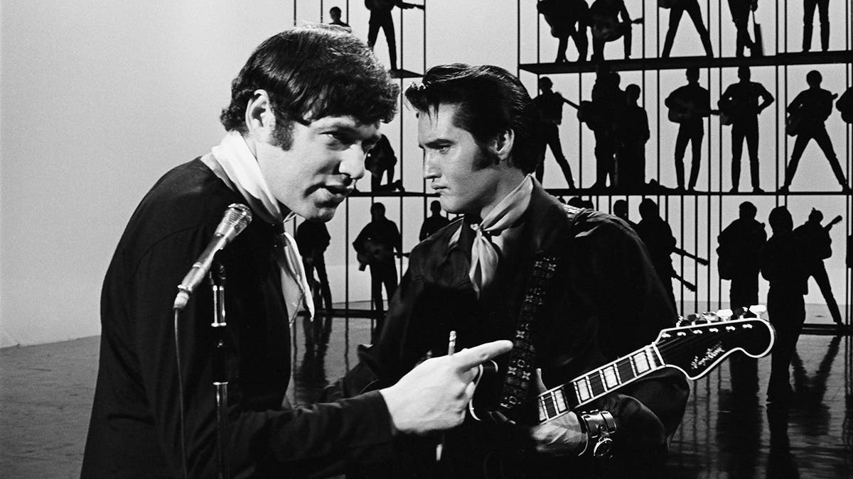 Steve Binder pointing in front of a camera as Elvis Presley looks away holding a guitar