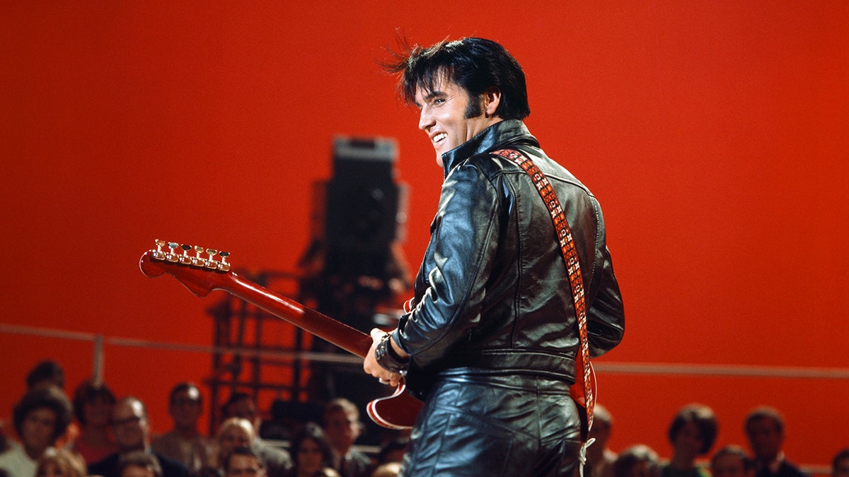 Elvis Presley looking from behind wearing a leather suit and holding a red guitar