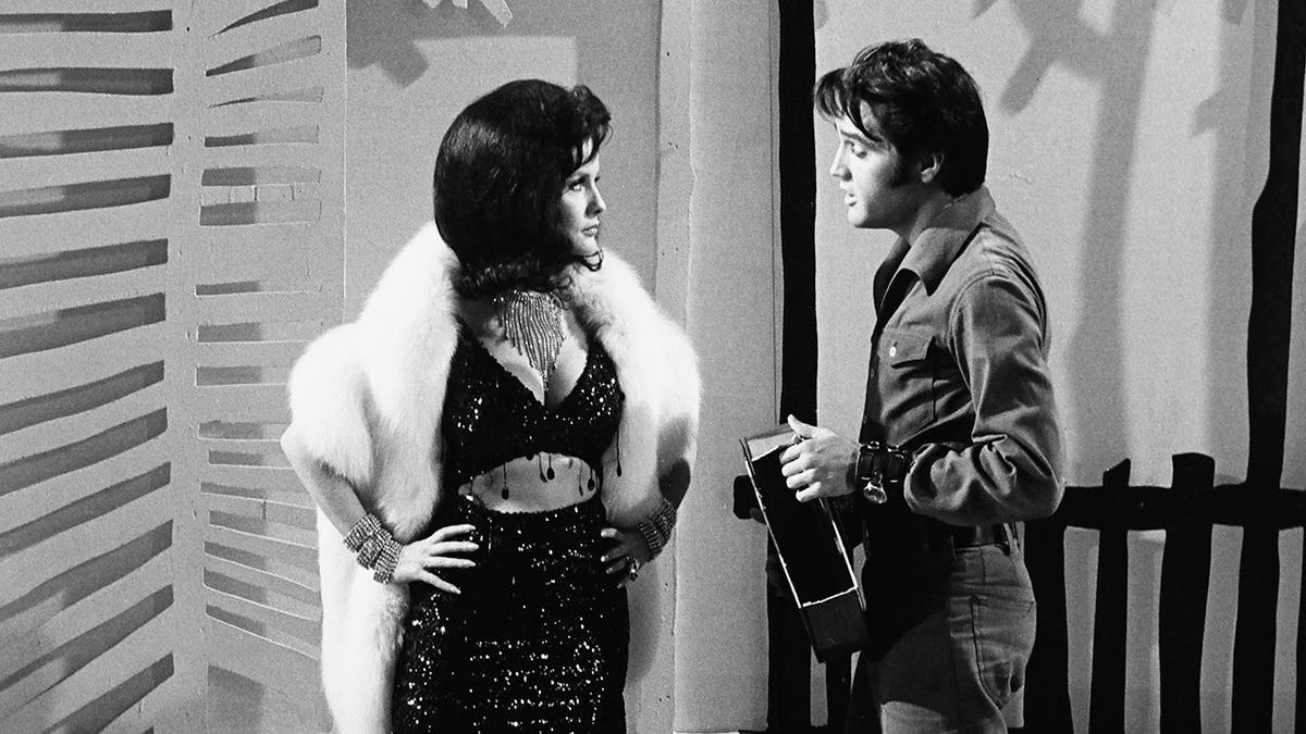 A model wearing a bejeweled bra top and skirt in a white fur coat talking to Elvis Presley in a denim suit