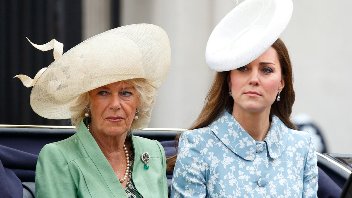 Queen Camilla wearing a green dress and an ivory hat sitting next to Kate Middleton in a blue and white floral dress and white hat