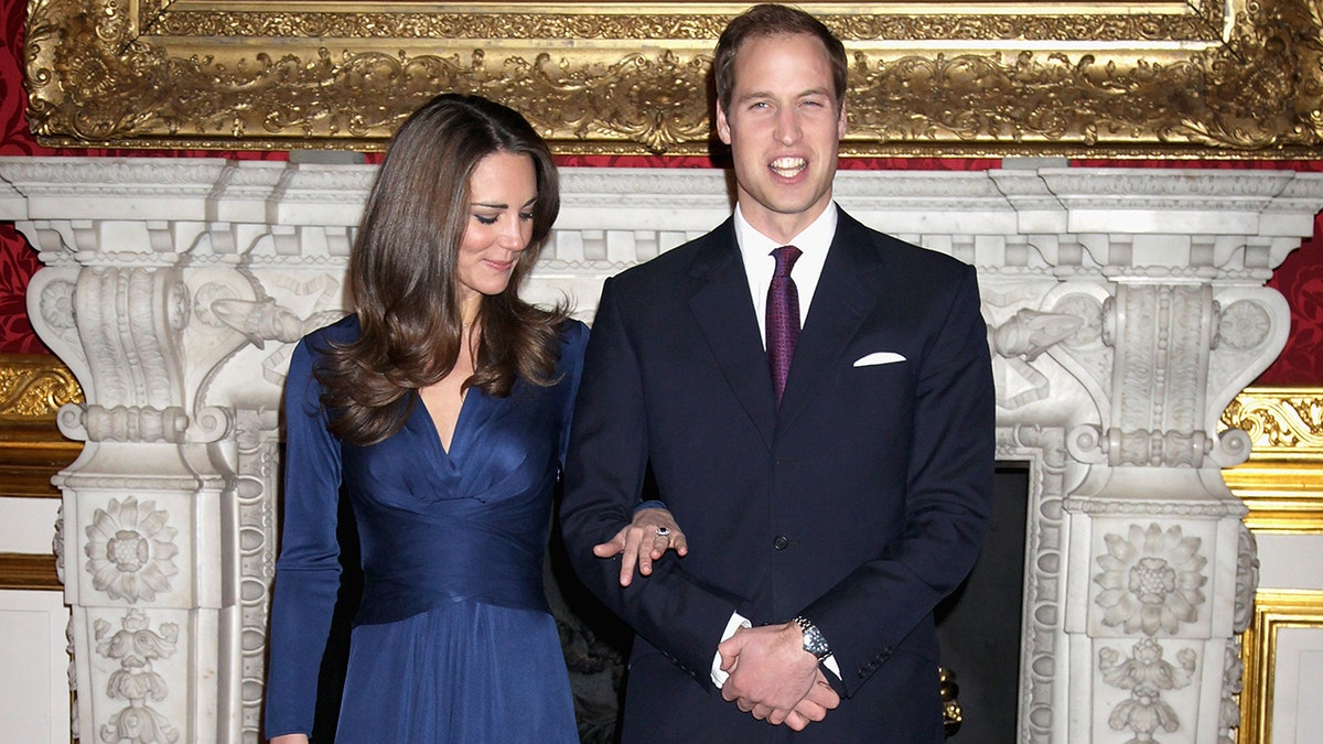 Kate Middleton in a blue wrap dress staring at her engagement in the arm of Prince William