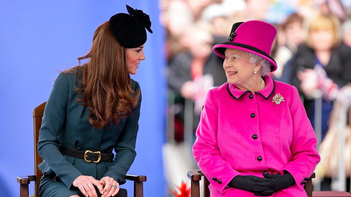 Kate Middleton in a hunter green dress and black fascinator chatting with Queen Elizabeth wearing a hot pink dress coat and matching hat