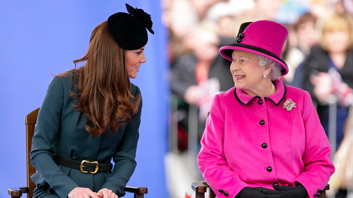 Kate Middleton wearing a hunter green dress sitting next to Queen Elizabeth wearing a bright pink dress