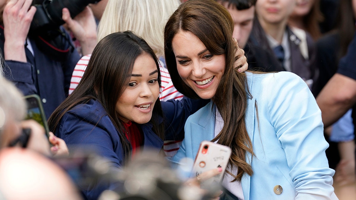 Kate Middleton in a light blue blazer and white blouse taking a selfie with a woman in a navy blazer