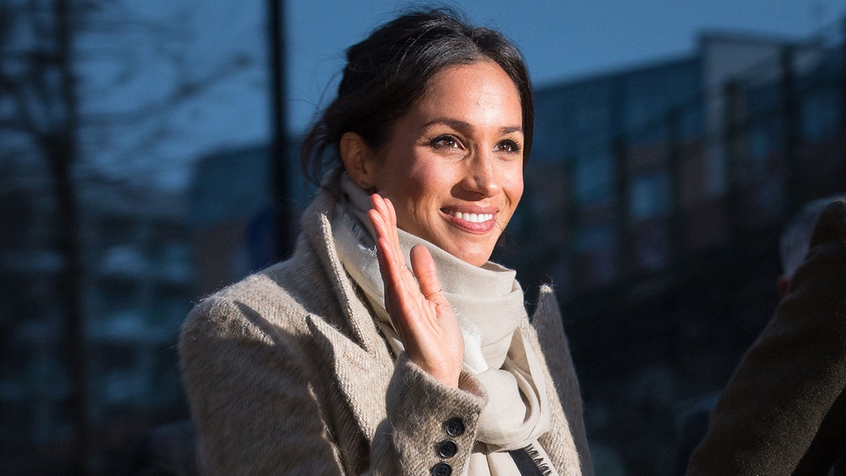 Meghan Markle waving and wearing a beige coat with a matching turtleneck sweater