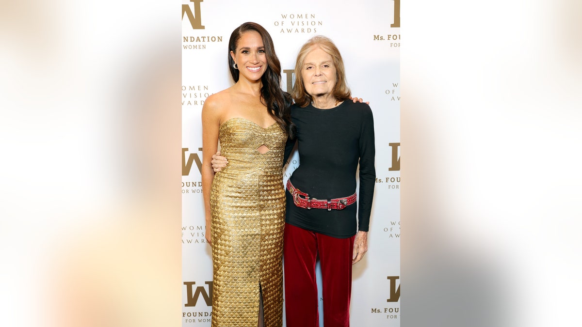 Meghan Markle in a gold strapless dress posing with Gloria Steinem who is wearing a black long sleeved shirt and burgundy pants