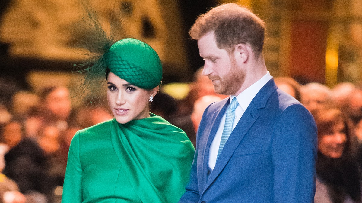 A close-up of Meghan Markle wearing a green dress and a matching hat standing next to Prince Harry in a blue suit and tie