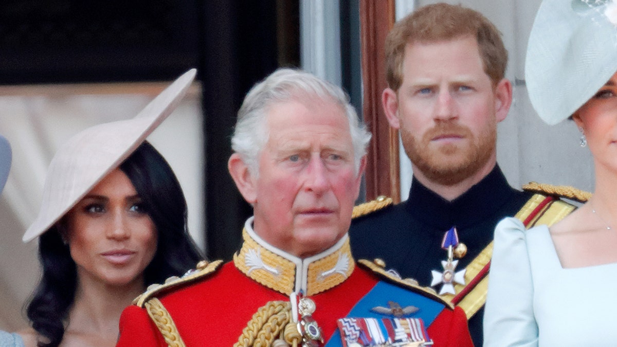 King Charles in a red military uniform in front of Prince Harry in a military uniform and Meghan Markle in a pale pink dress and matching hat
