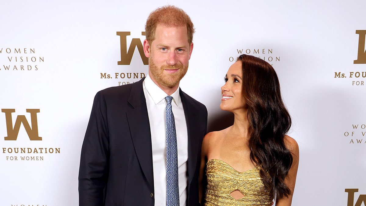 Prince Harry wearing a navy suit and a white shirt with a light blue tie posing next to Meghan Markle wearing a strapless gold dress