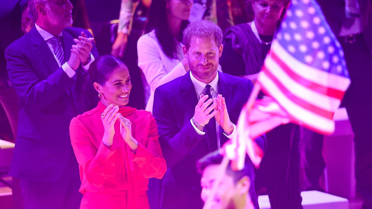 Meghan Markle wearing a red jumpsuit and Prince Harry wearing a blue suit in front of the American flag