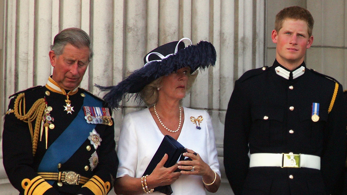 Prince Harry in a military suit looking annoyed as Camilla wears a white dress and an oversized navy hat standing next to prince charles in a military uniform