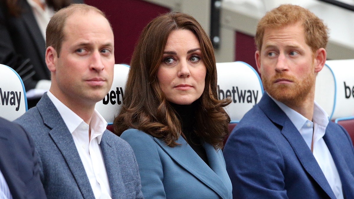 Kate Middleton wearing a blue blazer with a black sweater sitting in between Prince William and Prince Harry wearing various shades of blue and grey