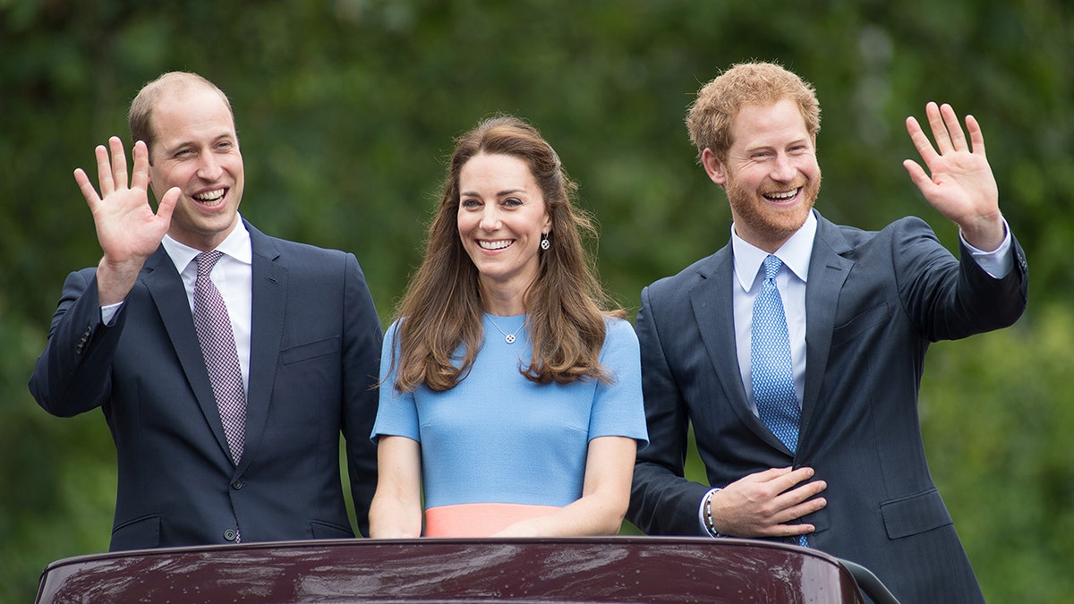 Kate Middleton wearng a light blue dress with a salmon-hued belt standing in between a smiling prince william and prince harry