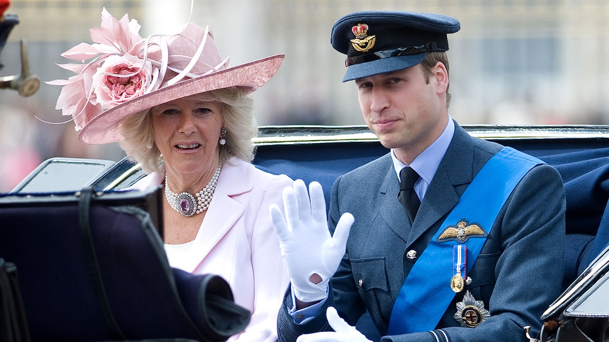 Prince William in a military suit sitting next to Queen Camilla in a pink dress and matching hat
