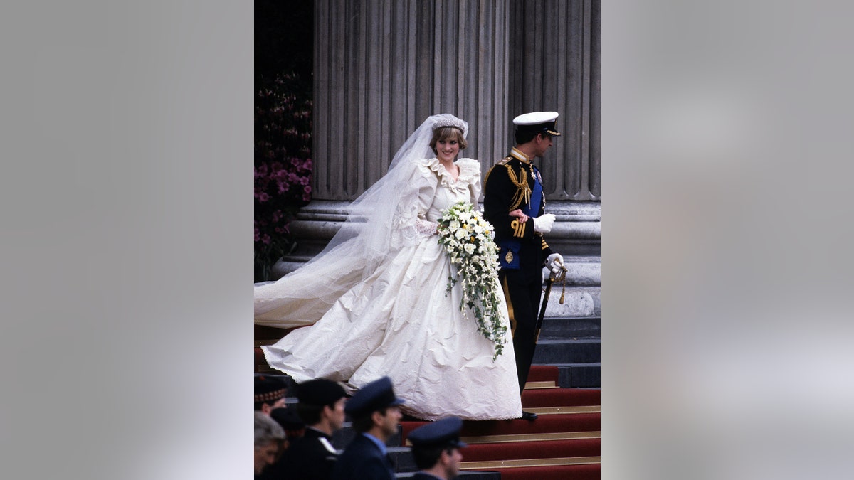 Princess Diana smiling in her wedding gown while holding a bouquet of flowers and walking alonhside Prince Charles