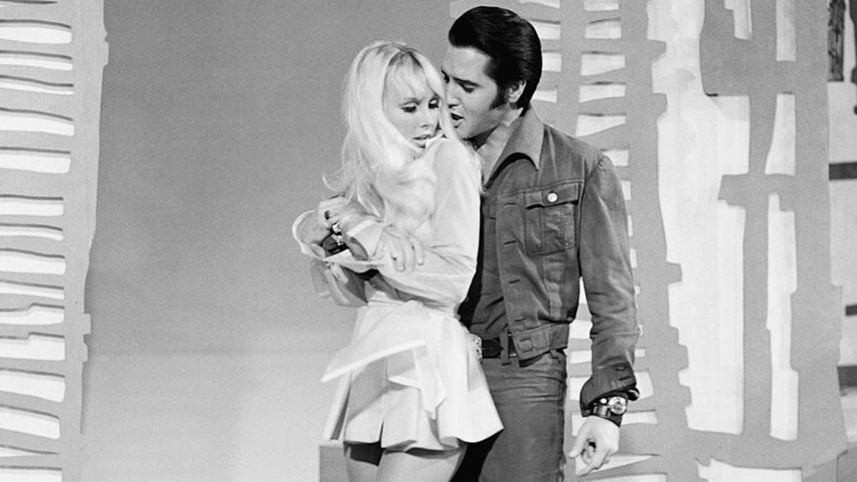 Susan Henning in a tiny white dress being held by Elvis Presley in a denim suit