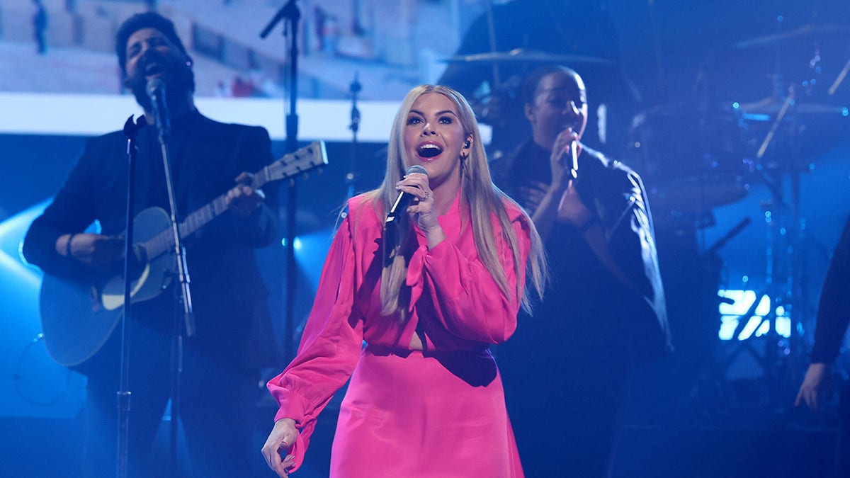 Tasha Layton wearing a hot pink jumpsuit and singing on a mic