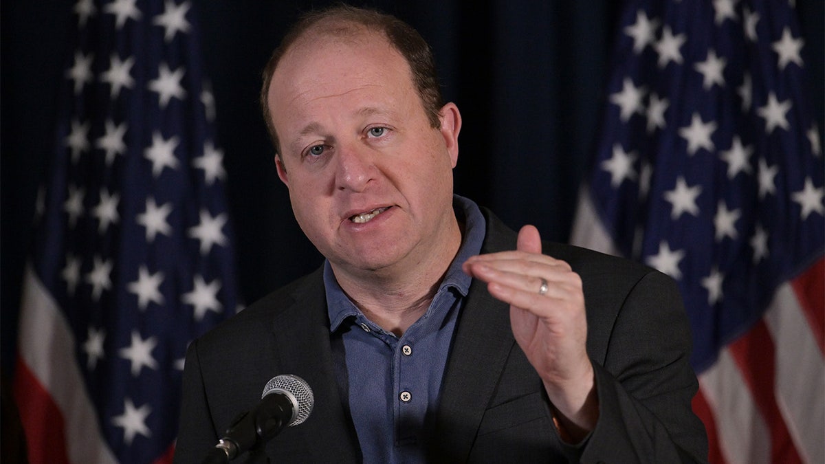 Jared Polis speaking with American flags in the background