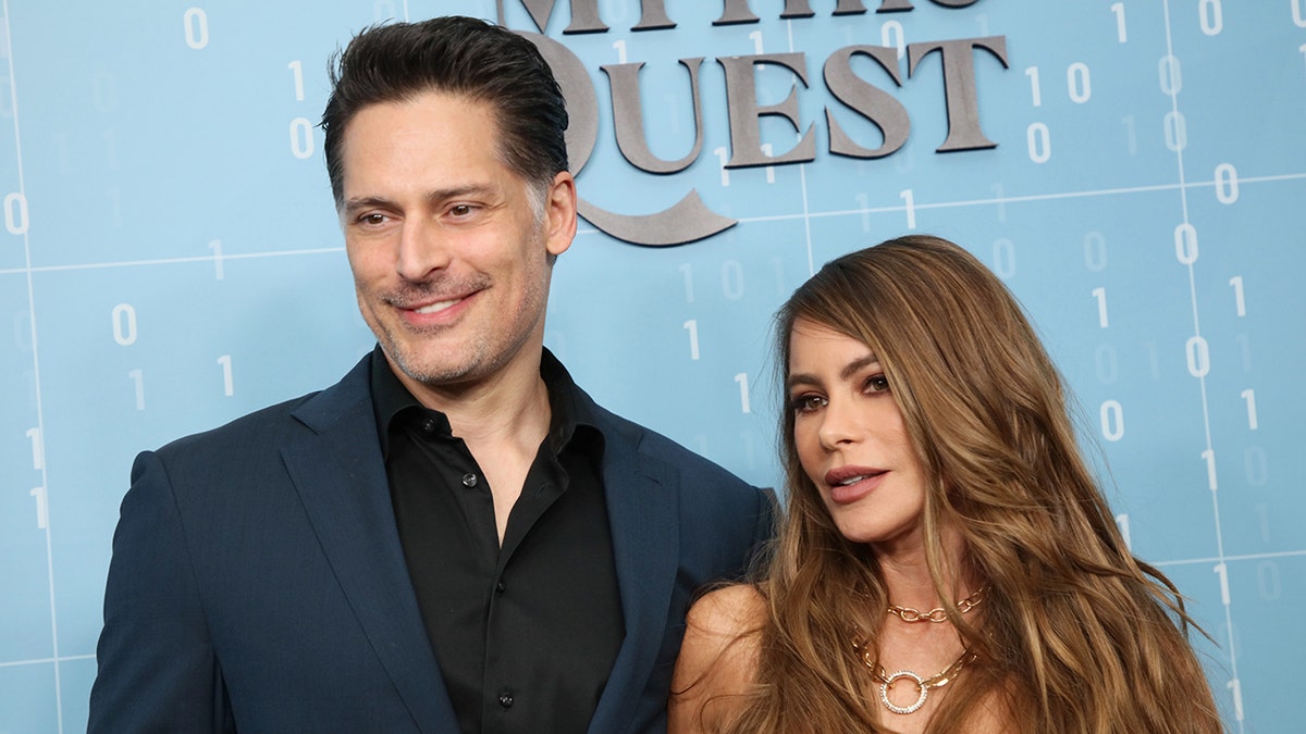 Joe Manganiello in a navy blue suit and black shirt poses with Sofía Vergara on the carpet